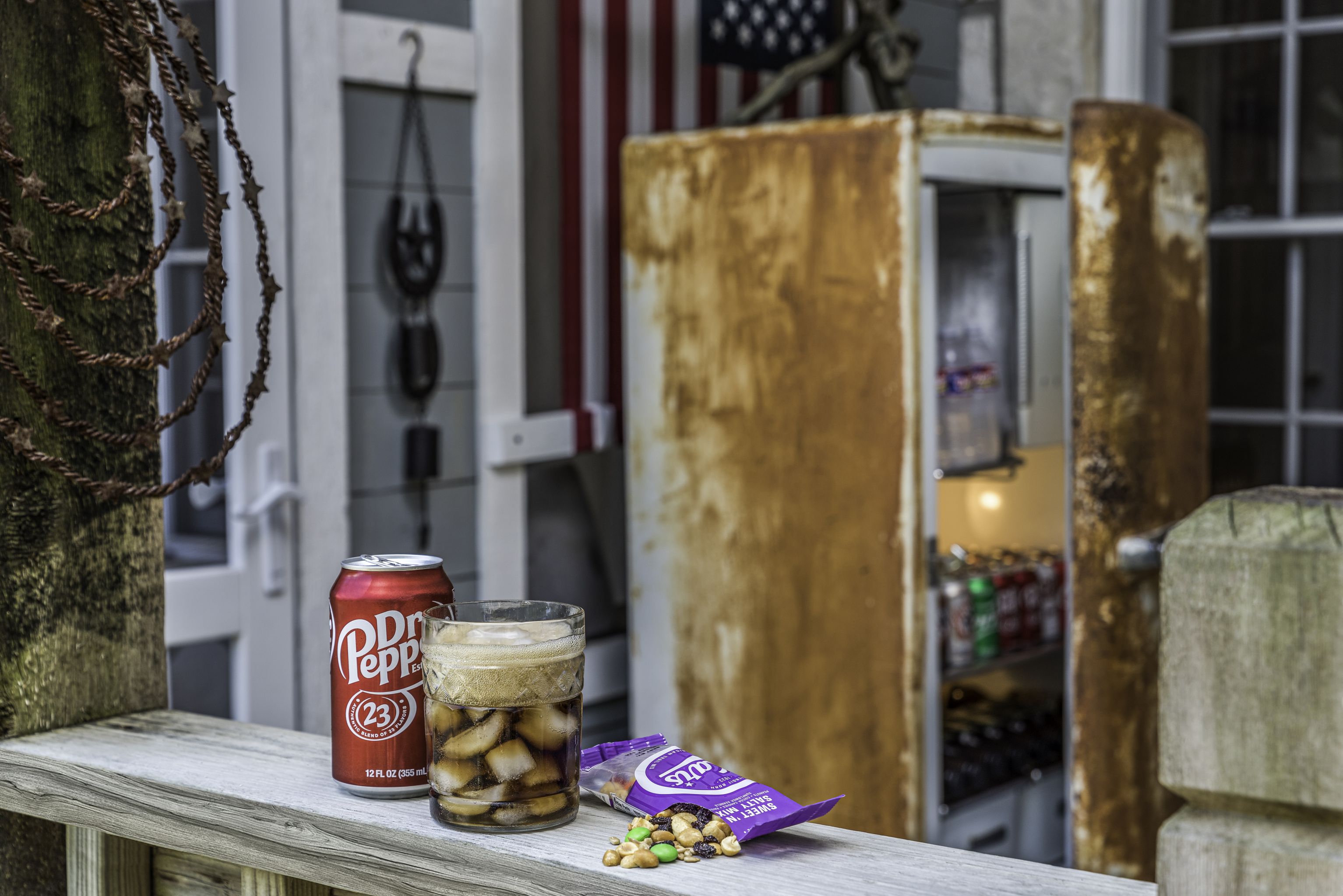 A vintage soda machine sits on a porch, with a can of soda, a glass of soda, and a bag of snacks in front of it.