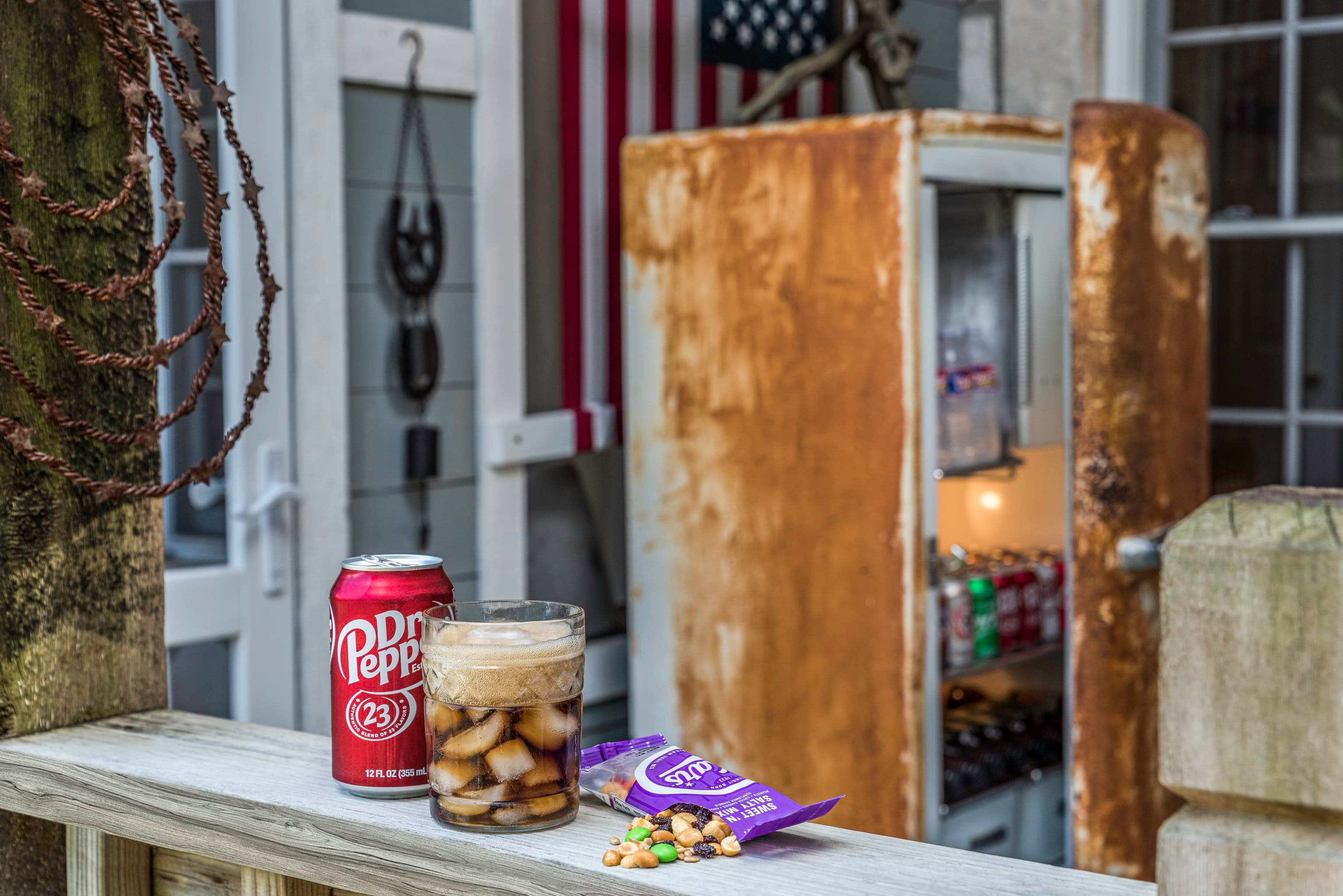A vintage soda machine sits on a porch, with a can of soda, a glass of soda, and a bag of snacks in front of it.