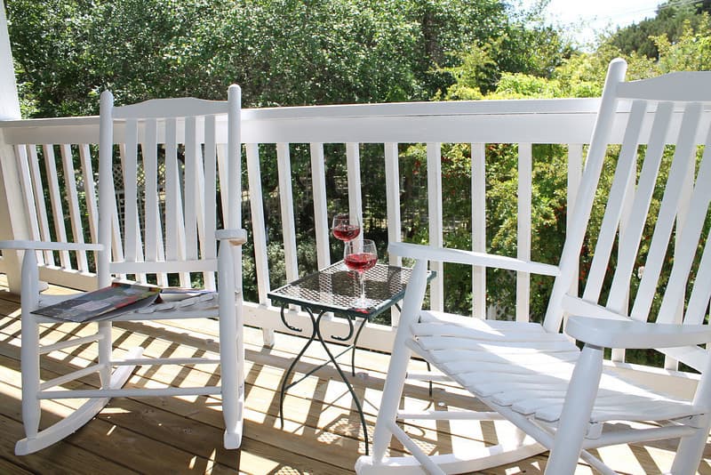 A view of a porch with two white rocking chairs and a small table holding a glass of wine. The porch has a white railing and overlooks a green, wooded area.