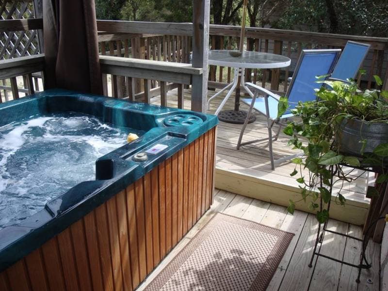 A hot tub on a wooden deck with a view of the woods. There are chairs and a small table nearby.