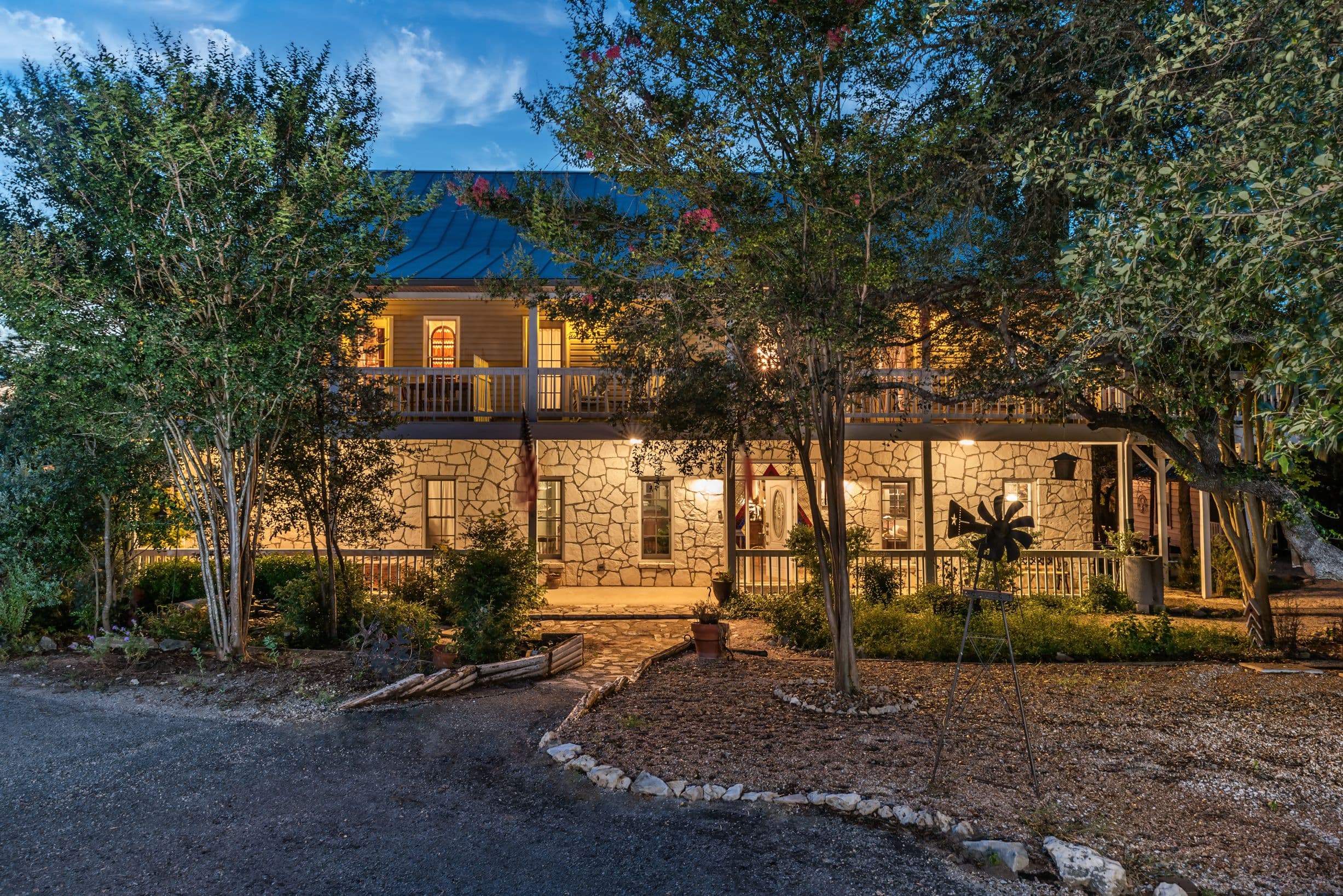 A large, two-story stone house with a wraparound porch. The house is lit up at night, and there's a path leading up to the front door. Trees and bushes surround the house.