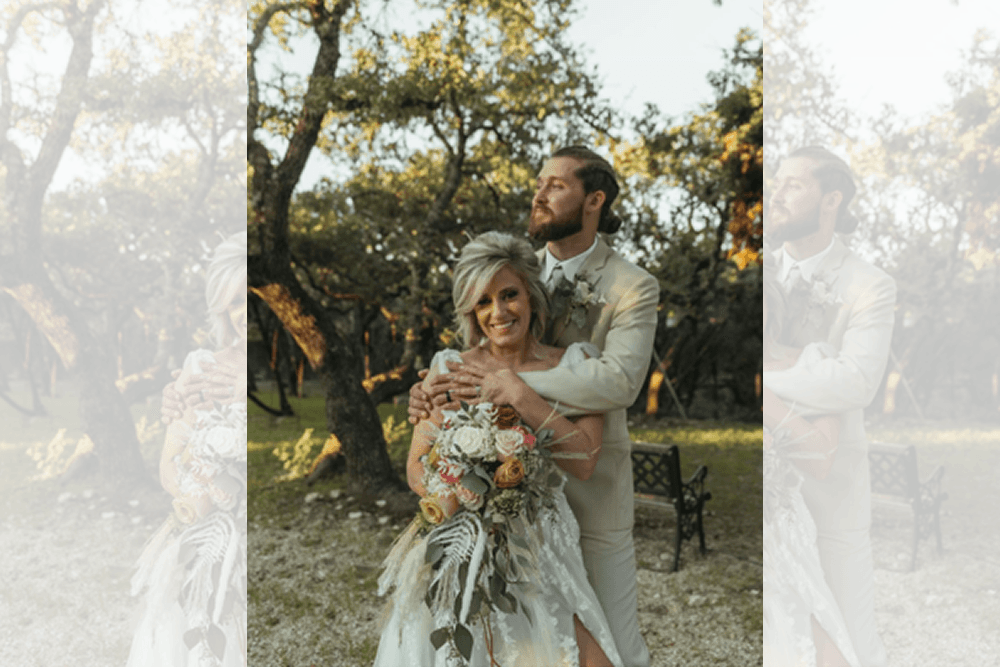 A bride and groom embrace outdoors, surrounded by trees and natural light.