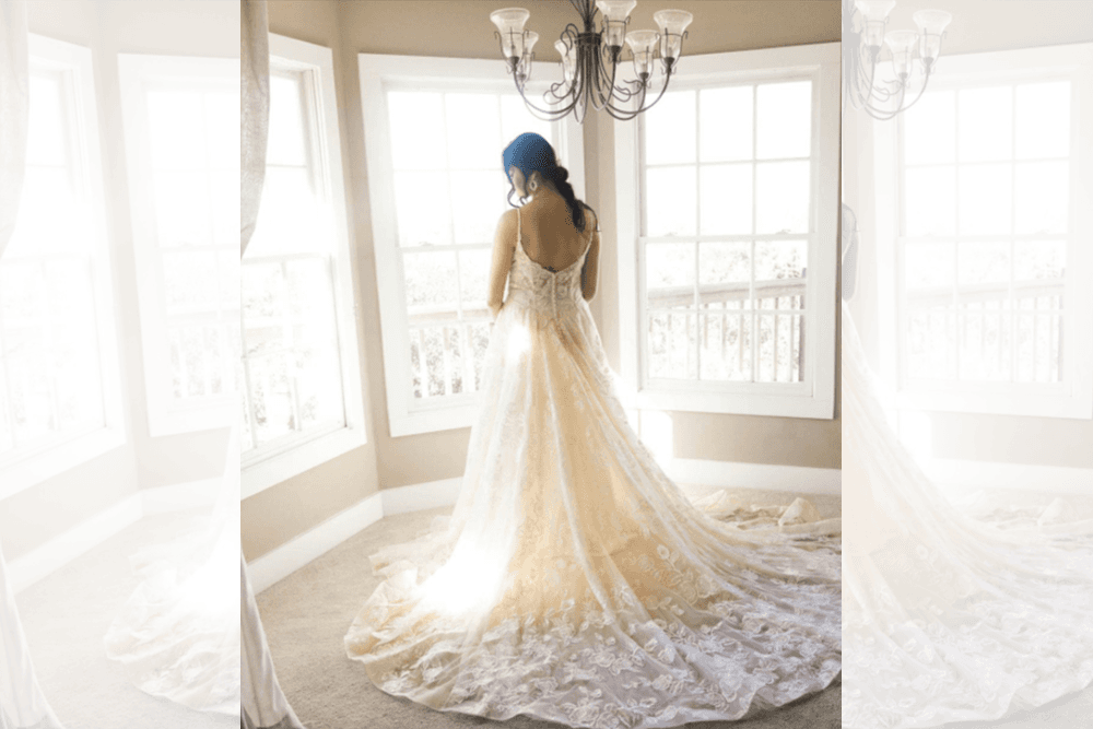 A bride in a stunning lace dress gazes out a window, with sunlight illuminating her train.