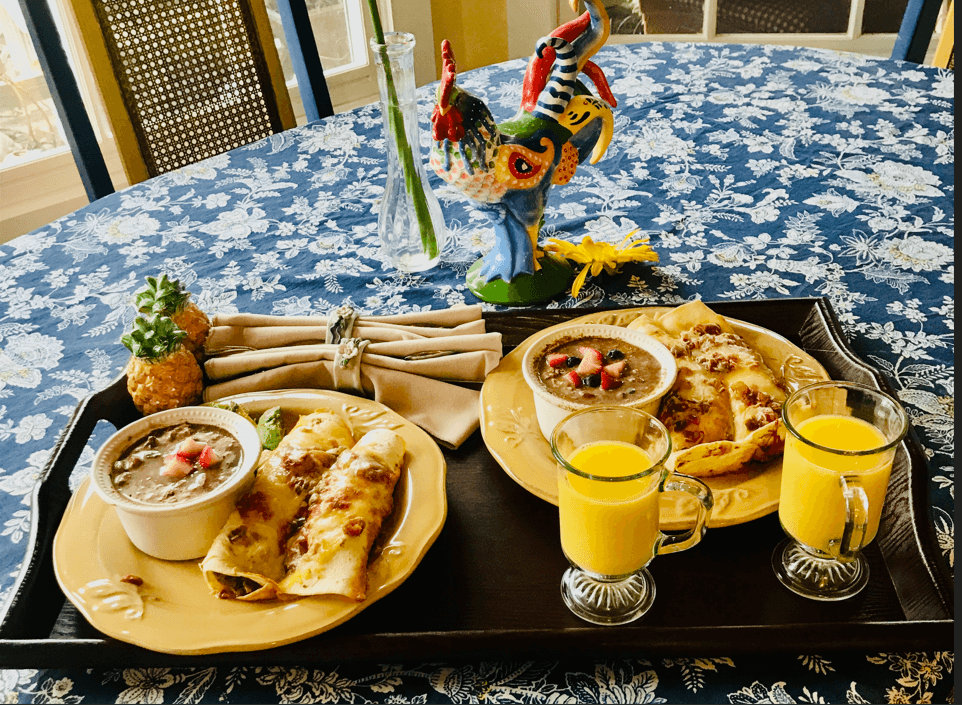 A vibrant breakfast spread featuring enchiladas, beans, and two cups of orange juice, set on a floral tablecloth with decorative elements.
