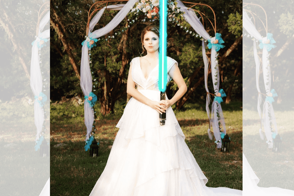 A bride in a flowing white wedding gown stands with a glowing blue lightsaber in front of a decorated arch.