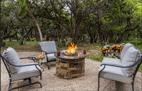 A cozy outdoor seating area with a fire pit, surrounded by greenery and logs.