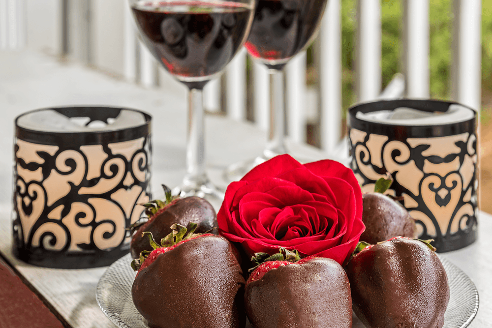 A romantic arrangement featuring chocolate-covered strawberries, a red rose, and two glasses of red wine beside decorative candles.