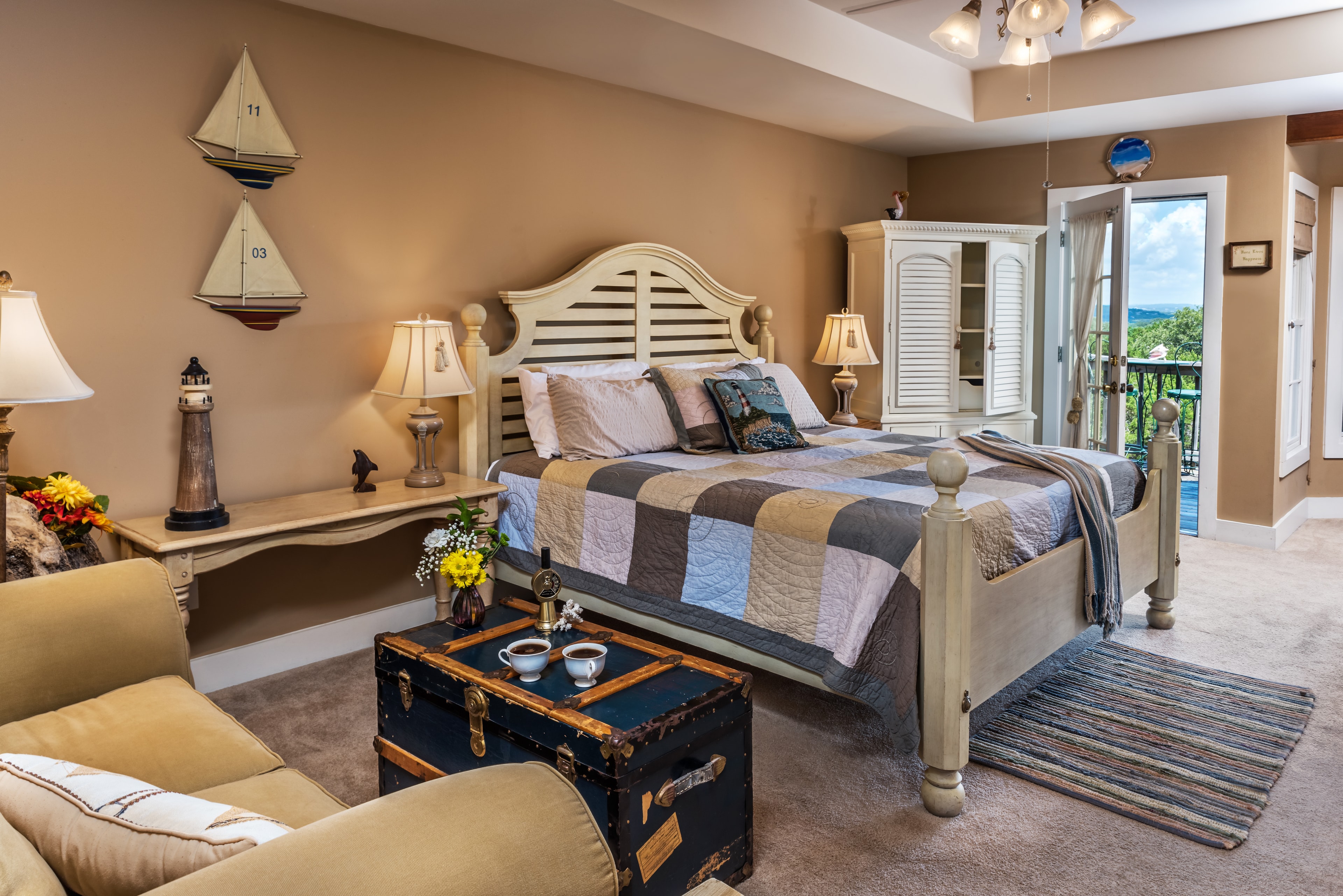 A bedroom decorated in a nautical theme. The bed has a patchwork quilt and there's an armchair nearby. There's a table with a vase of flowers, and a large window with a view of the outdoors.