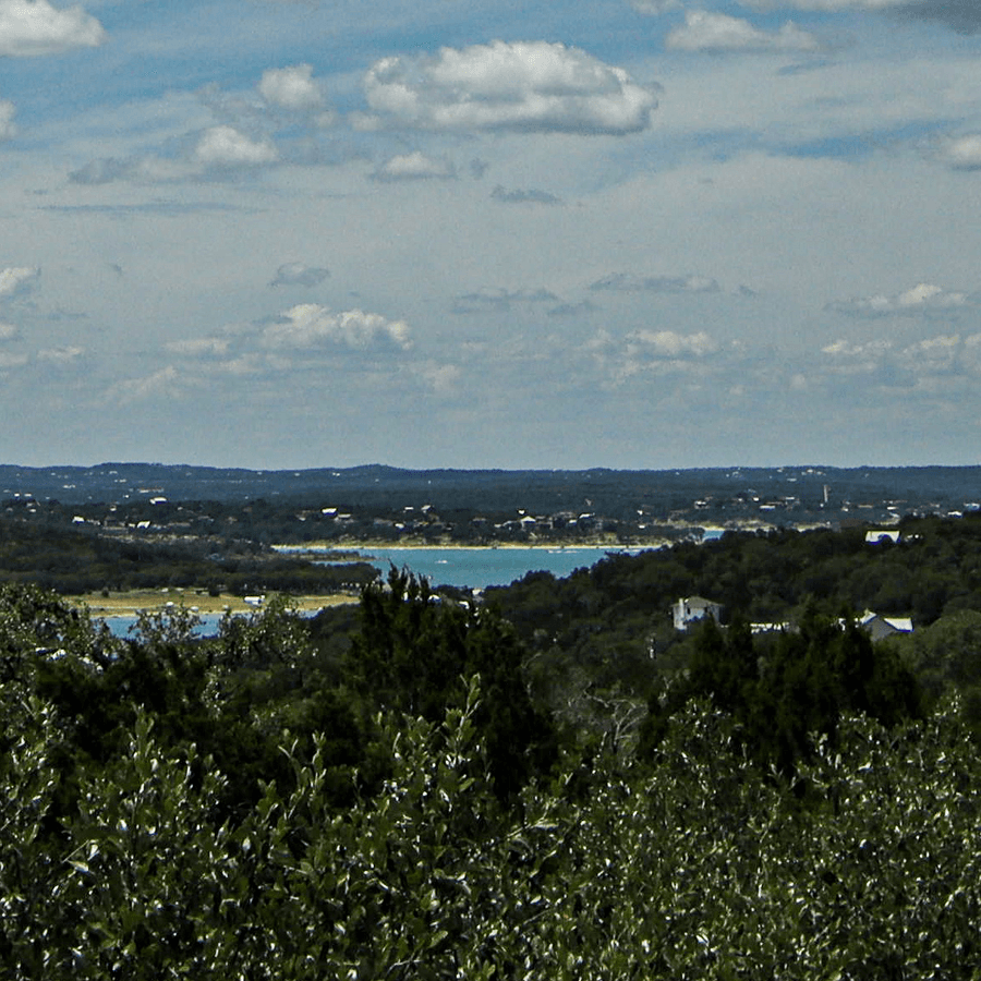 A panoramic view of a lake surrounded by greenery and distant hills under a cloudy sky.