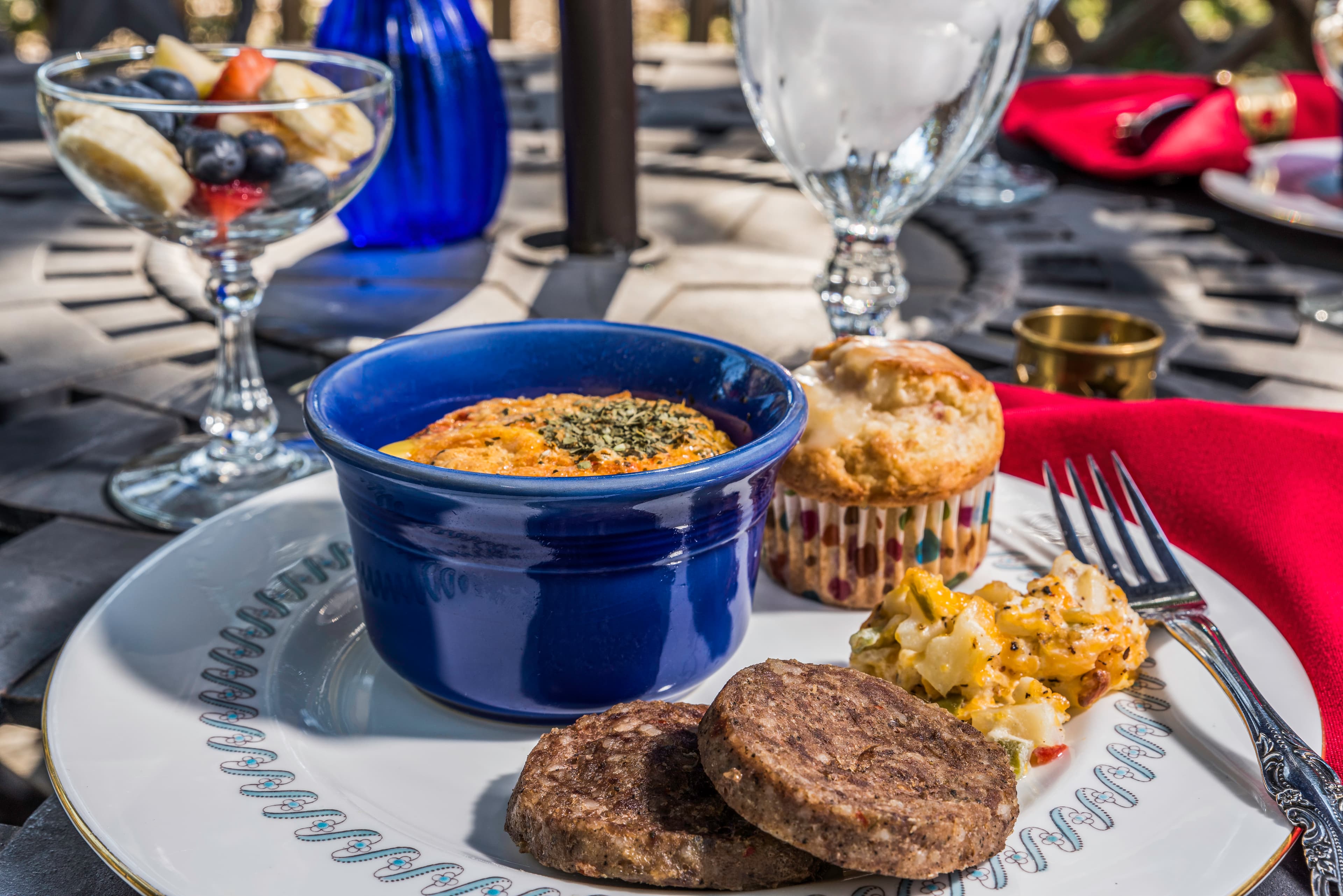 A plate featuring a baked dish in a blue ramekin, two sausage patties, a muffin, and a side of macaroni, accompanied by a fruit salad in a clear bowl.